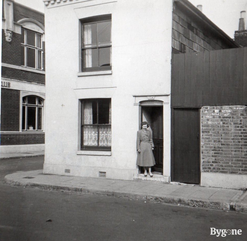 There is a house at the end of a street with a door slightly ajar and a woman standing outside in front of it. The woman is wearing a long double breasted coat with a belt and her hair is in short curls.