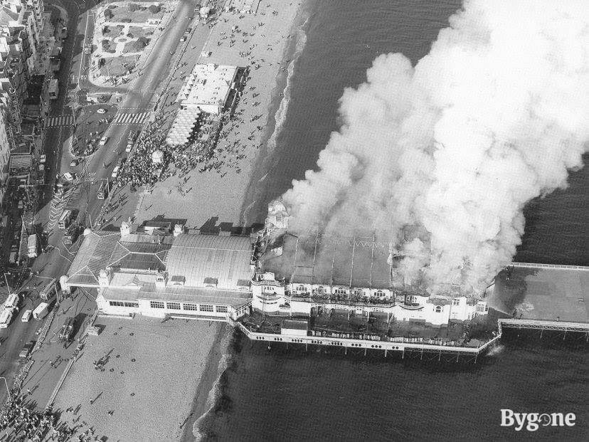 View of a pier in flames from above. Tumultuous clouds of smoke swirl upward from a long building raised above the sea. Swarms of people are watching the blazing fire unfold nearby.