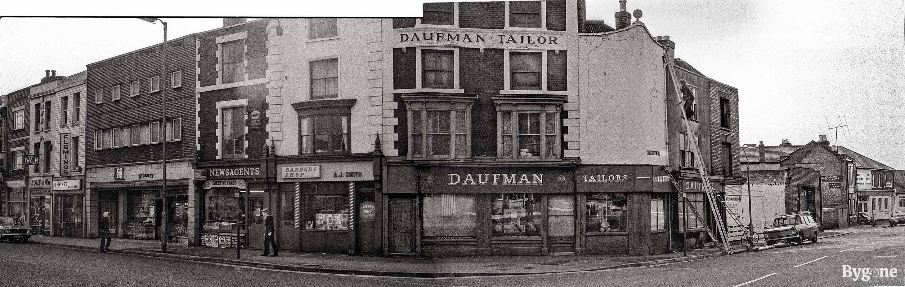 Panorama of a street corner with shops. From left to centre right the shops are, Baun & Co, Flemings, Coop grocery, Newsagents, Barbers Shop A.J. Smith, and Daufman Tailors. To the right there is a man climbing up a tall ladder to the side of Daufman Tailors.