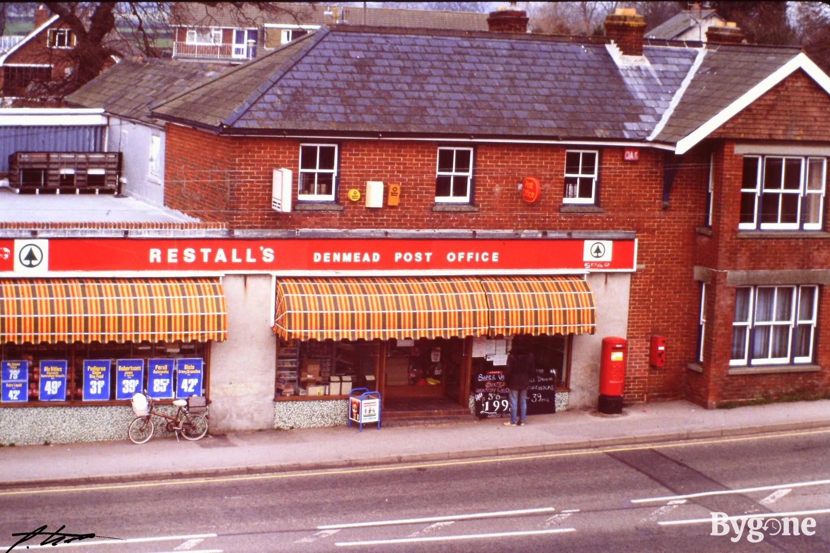 A vivid street shopfront with a red and white sign, above yellow and brown check canopies, that reads Restall’s Denmead Post Office. Spar. A man is standing outside looking at the advertisements stuck in the window.
