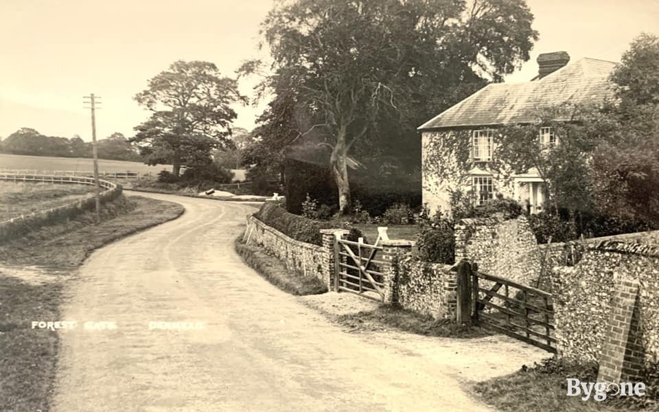 A winding road in a rural area, that passes by a large house with ivy growing up its front. To the right there is a gate by a stone wall with a path.