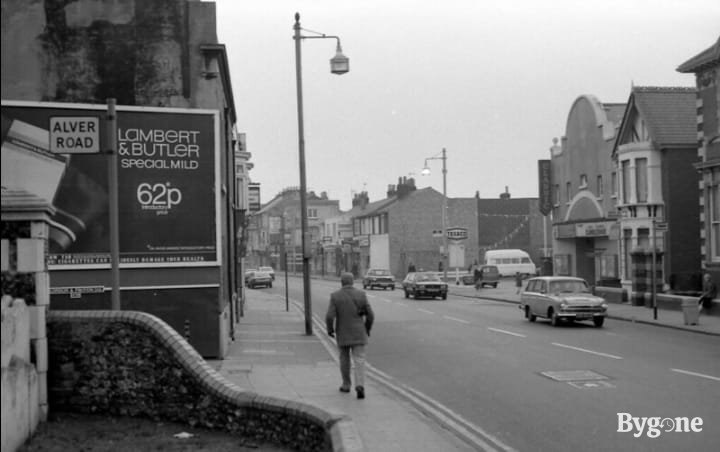 Street view with cars passing, and an unknown man walking up the street. There is a billboard to the left that says Lambert & Butler special mild 62p. To the left there’s the start of an alleyway with a street sign saying Alver Road.