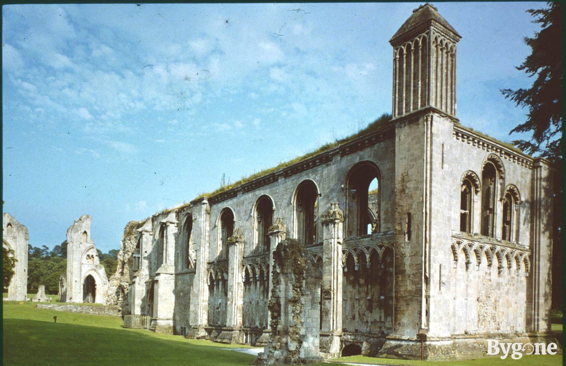 Beautiful stone ruins of an abbey in a green field on a sunny day, with arched windows and mossy grass growing on parts of its roof. A low wall leads to more monolith ruins with arches standing nearby in the distance.