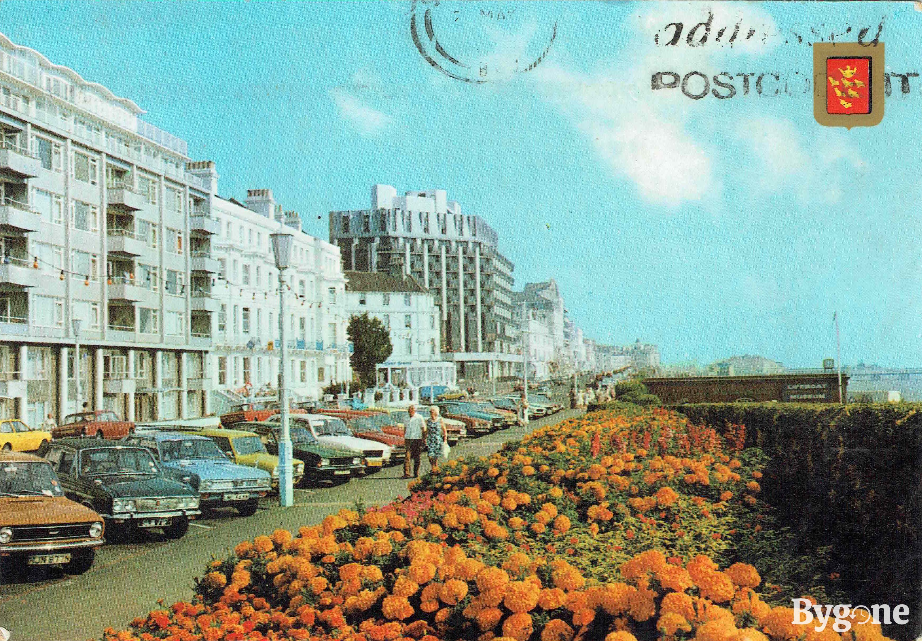A white middle-aged couple are walking hand in hand along a seafront parade. There are flats, houses and tall buildings along the left hand side, and a row of colourful cars parked diagonally. Alongside the pavement to the centre right, a flower bed of glorious orange marigolds are blooming, and the sky is painted a soft pale blue.