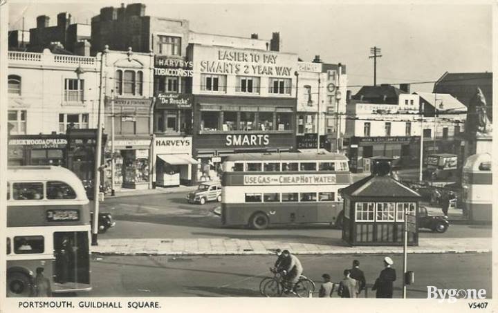 A busy street square with a strip of shops, pubs and hotels, some of the signs read: Harvey’s Tobacconists. Smarts. Chemists. Hotel. In the road double-decker buses, cars and bicycles are whizzing by.
