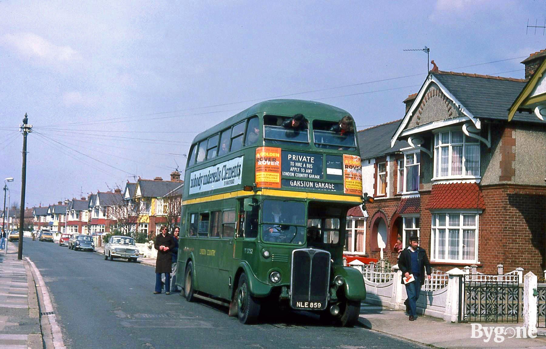 A green double decker is driving down a street with a private party of people on board. There are two people poking their heads out of the front windows on the top deck.