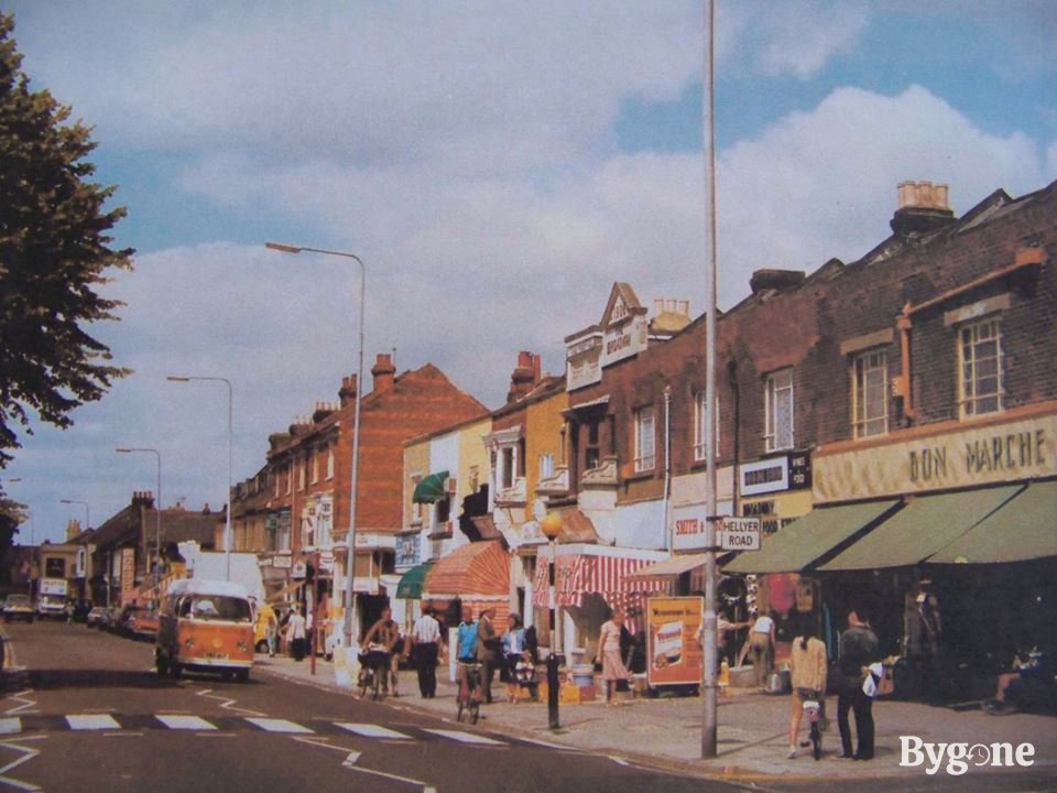 A sunny day with crisp clouds on a street corner. Many people are out and about, looking at and walking by a row of vibrantly coloured shop fronts with striped canopies, the shop sign on the right corner reads Bon Marche. An orange camper van is coming along the street by a zebra crossing.