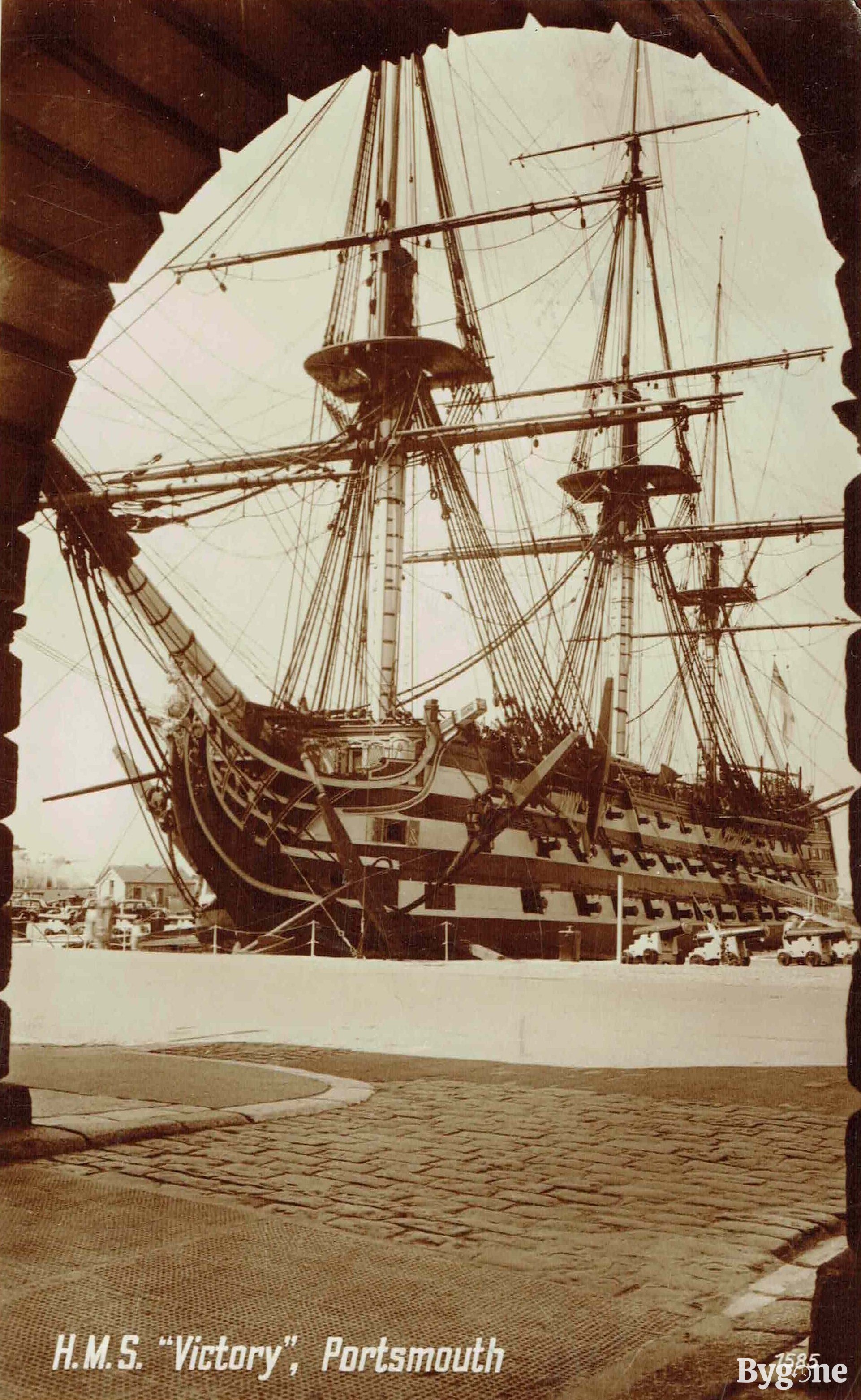 Portrait taken from underneath a stone archway of a large, three-masted, full-rigged ship, that is docked. The lower left reads 'H.M.S. "Victory", Portsmouth'