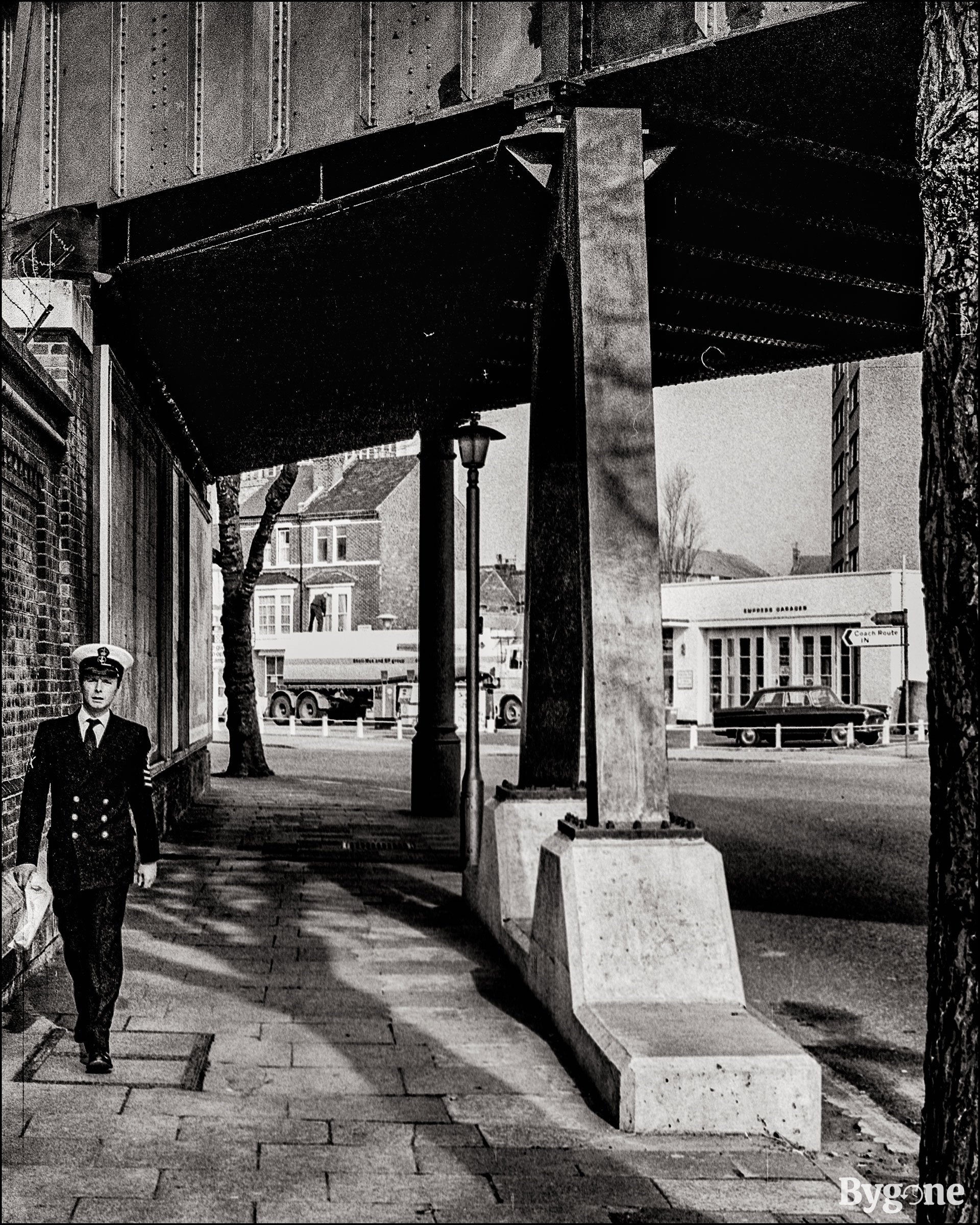 Street view from under an overhead railway bridge. There is an unknown man walking towards the camera on the left hand side of the frame, he is dressed in an old naval uniform. To the left there is a big pillar fixed into a stone plinth in the pavement. In the background there is a lorry and a car parked outside of a garage called Empress Garages.