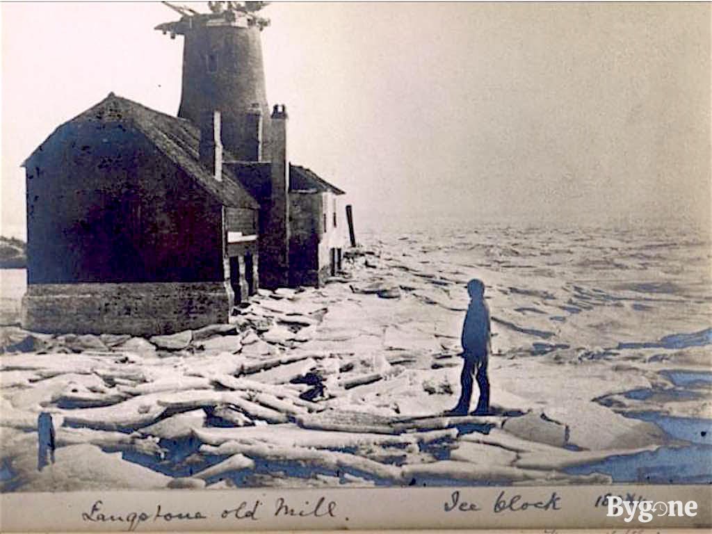 A lone man stands on large sheets of broken ice. He is looking toward an old mill that is blocked in, surrounded by ice.