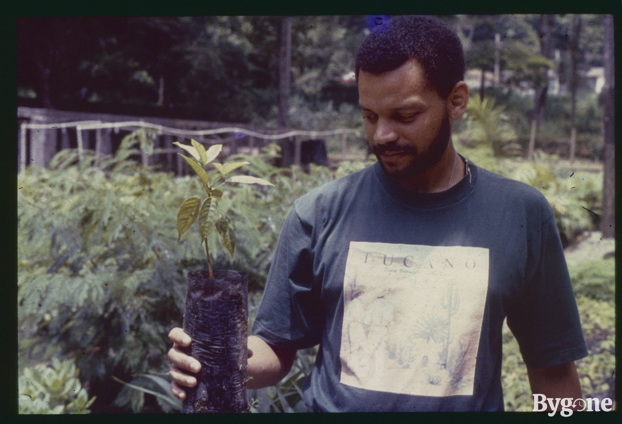 A young Black man with short dark afro hair and a close beard, is standing in a garden. He is wearing a shirt that reads Tucano, and is holding in his right hand a tree sapling. There are a handful of little leaves growing and its roots are wrapped in a thin sheet of plastic.