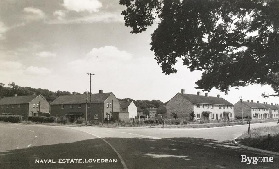 A wide road is veering to the left and has come to a junction on the right. A group of semi-detached houses are clustered by the road in the centre of the frame, making up a large estate.