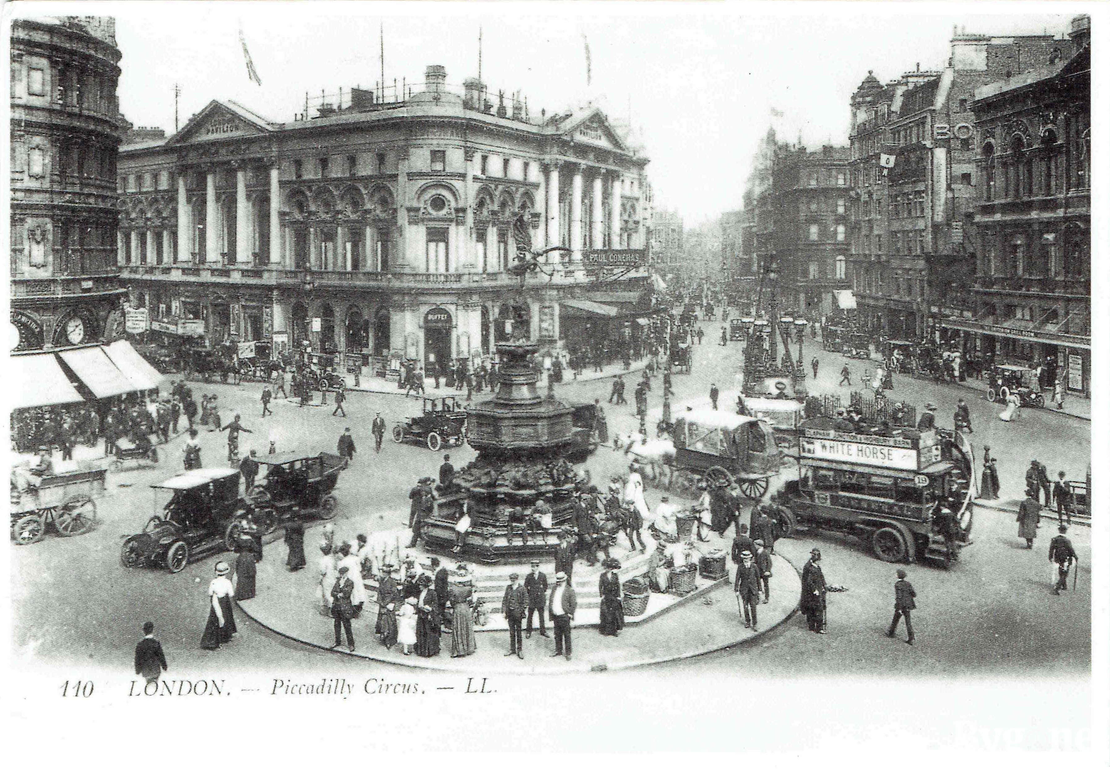 A bustling street junction, with a fountain surmounted by a winged statue in the centre of a roundabout. There are people sitting on the fountain's edge, women and men are standing by the steps, and walking along the pavements. Trams, horses pulling carts, and cars are passing by. The back of the postcard reads: ‘In this view c1908 the flower girls can be seen on the steps to the right of Eros’.