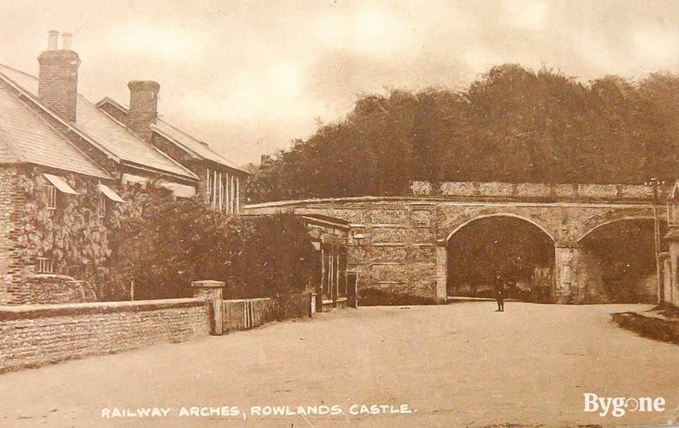 A group of houses sit on the left, their walls decorated with wisteria and ivy. Just to the right is a simple stone railway bridge with two arches beneath. In front of the left arch, one lone person stands in the middle of the empty street, their body but a speck, against the outline of the arch's grand void-like presence.