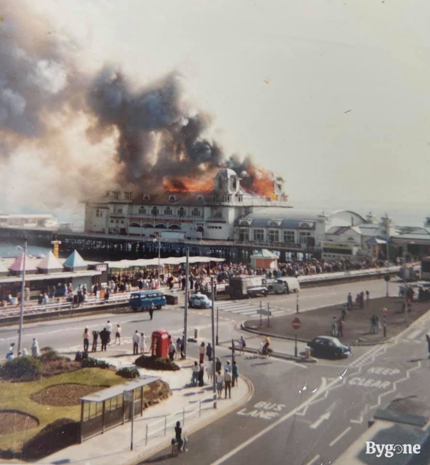 The roof of a pier is completely alight with orange flames, and grey clouds of smoke spew outward, tinged with an orange hue. Crowds of people are standing on the street and parade watching on as the building burns.