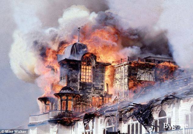 Close up of the roof of a building completely on fire. Hues of red-orange and golden flames engross the white wooden panels, turning them blue-black. Thick fumes, dark with grey and cloudy with white, undulate upward, pouring smog into the sky.