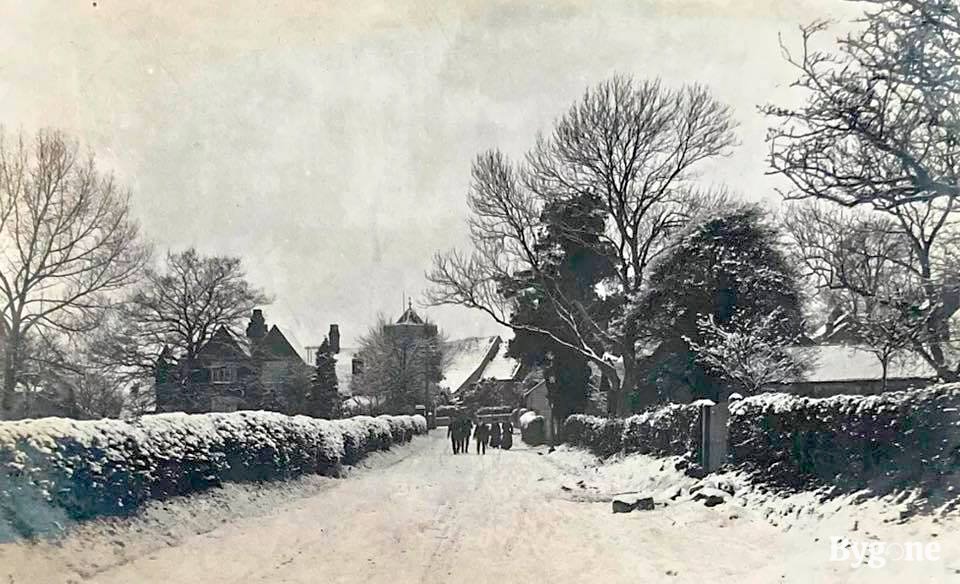 A group of people are walking together in the middle of a village road, which is caked in a thick white covering of snow. The tops of the hedgerows either side are dusted with ivory speckles. In the distance the roofs of the houses are white as sheets and pale strips of snow outline the branches of the surrounding trees.