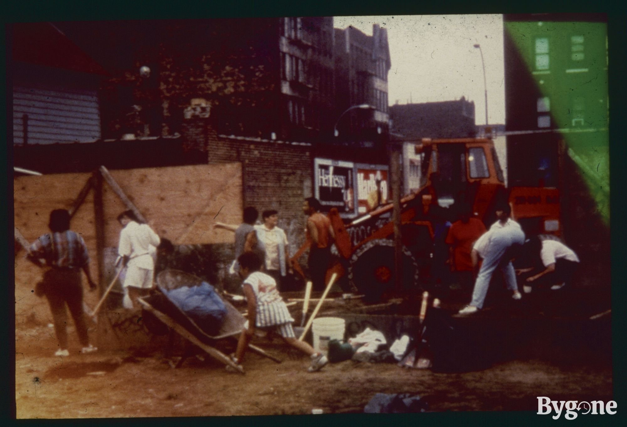 A group of people are standing and working on a building site, dressed in casual clothing. Some are in conversation, others crouched down in front of an orange tractor, whilst another is readying themselves to lift a wheelbarrow containing a heavy load. The dig is situated in a street with urban buildings surrounding it.