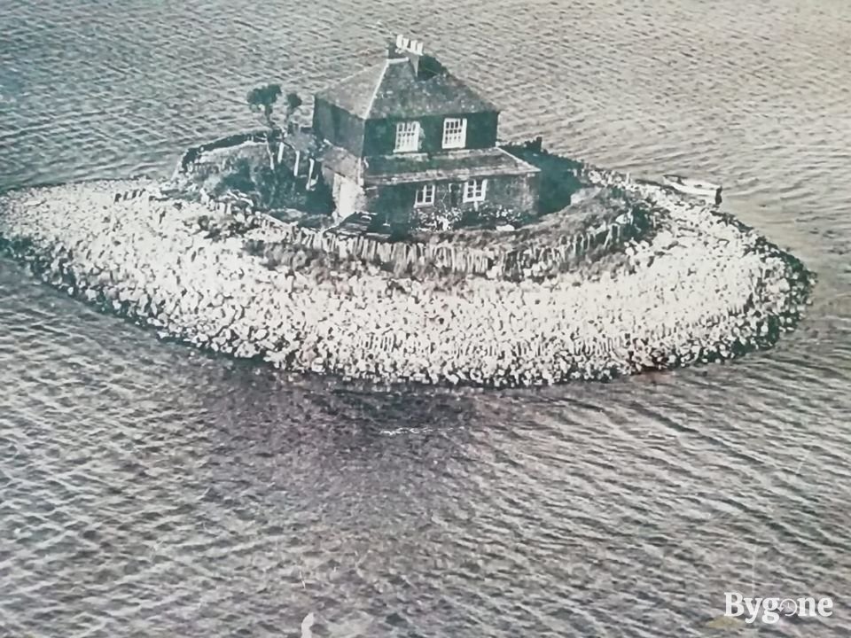 Aerial view of an ominous lodge whose walls are painted with black tar sits centrally on a small circular island of stones. The island is surrounded by soft rippling water.