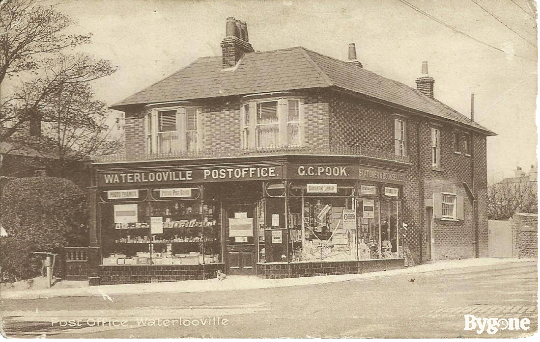 Landscape of a house with a post office shop front, and a little dog lying down outside the entrance door. The building’s frame reads, Waterlooville Post Office, G.C.Pook, Stationer & Bookseller.