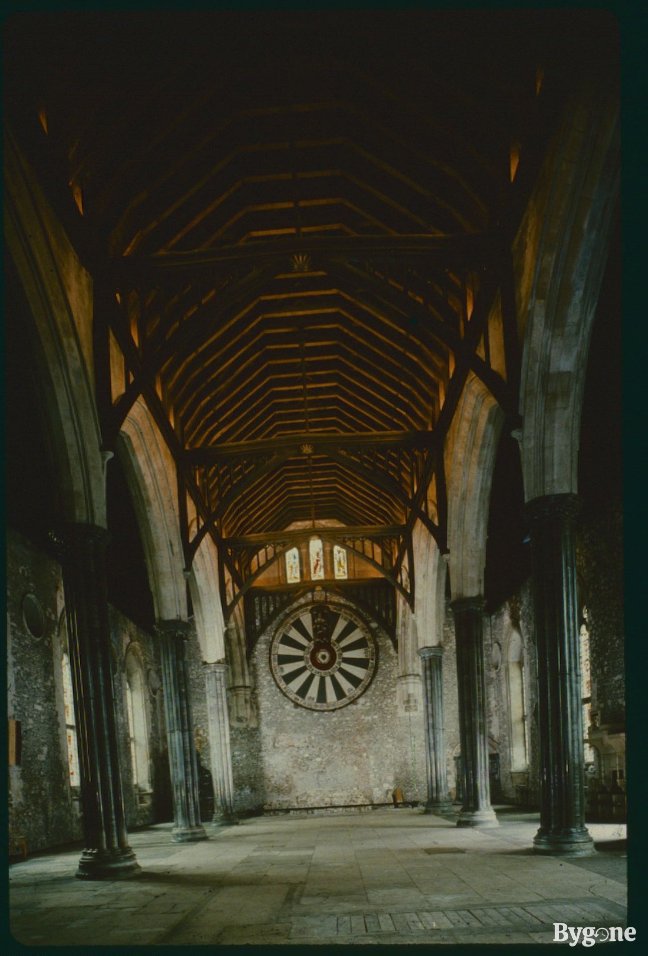Looking down the centre of a castle’s great hall toward a round black and white table that’s mounted into the centre of a stone wall at the end. There are wooden hexagonal arches overhead and black stone pillars with arches either side of the central walkway.