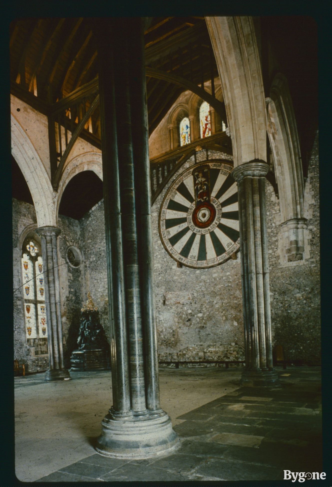 A section of a castle’s great hall. Taken from behind a stone pillar arch, looking up at a black and white round table that has been mounted into the centre of a stone wall. In the table’s centre is depicted a Tudor Rose; a double rose with white petals for the inner flower and red for the larger outer rose. Above the rose is depicted a seated King Arthur holding an upright sword.